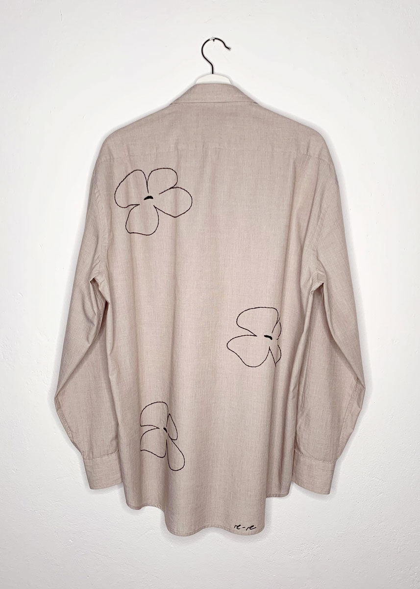 Beige shirt on a hanger from the back. Featuring 3 big hand embroidered flowers.